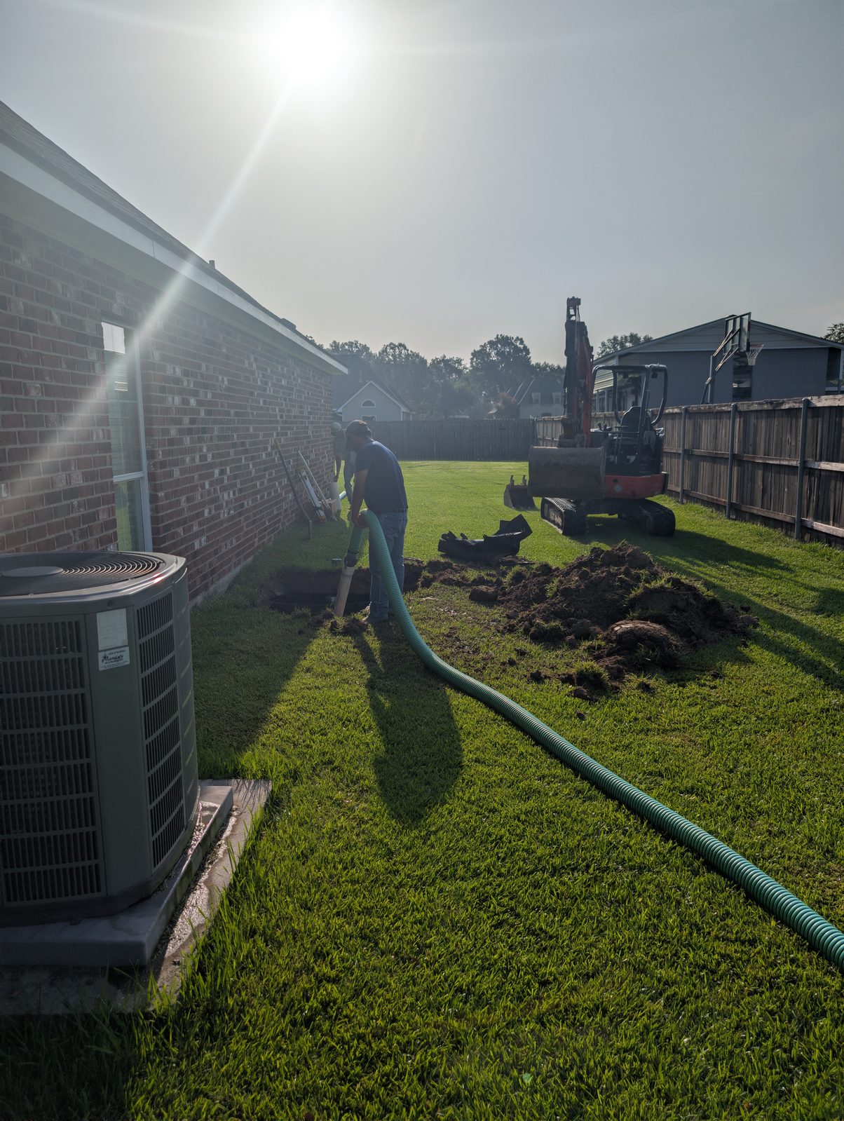A St. Amant Septic technician running the suction hose to a tank alongside a brick home, with the excavator standing by.