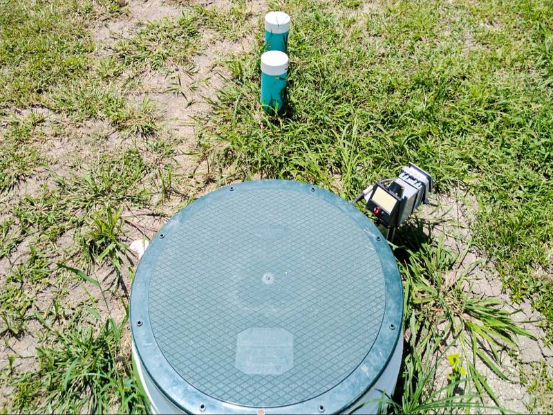 A finished green riser lid set flush in the lawn, with the control box beside it and two cleanout risers behind.