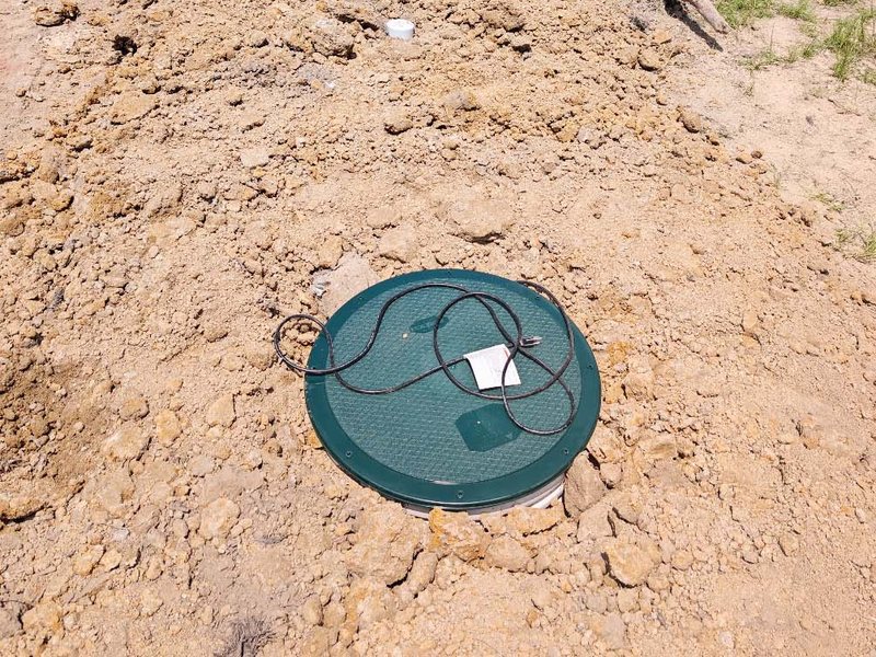 A green riser lid set in fresh backfill on an open lot, the pump power cord coiled on top.
