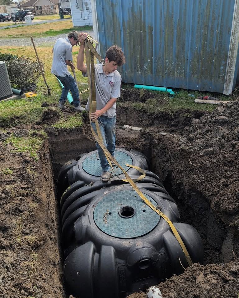 Two crew members guiding a tank down into its excavation on straps.