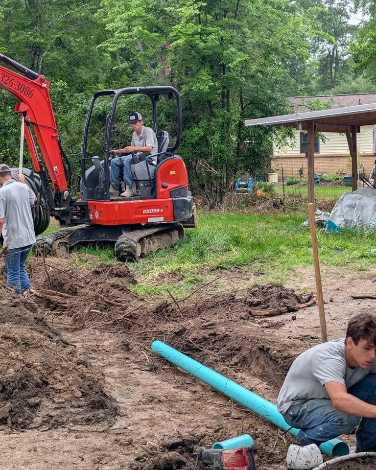 A crew member fitting new pipe while the excavator holds the trench open.